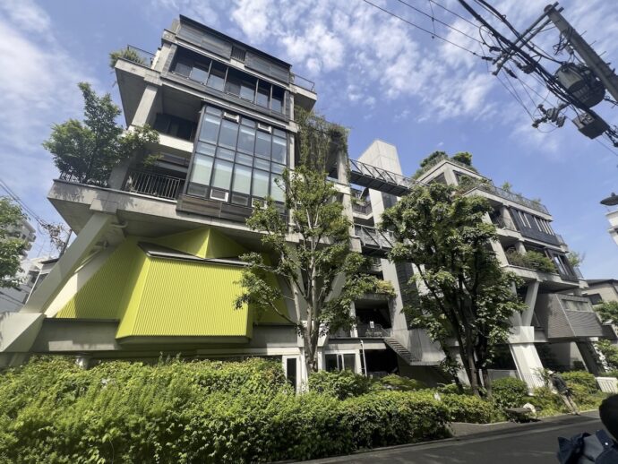 Modern concrete apartment building with cantilevered floors, large glass windows, and a bright lime-green corrugated wall panel surrounded by trees on a street.