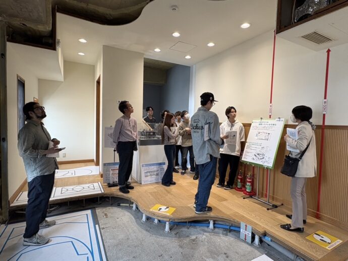 A group of people in a showroom listen to a presenter beside a large poster on a stand, with others looking on and a ramped wooden platform in the foreground.