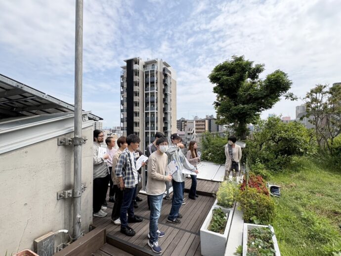 Group of people on a rooftop garden tour, holding papers and listening to a guide near a row of planters.