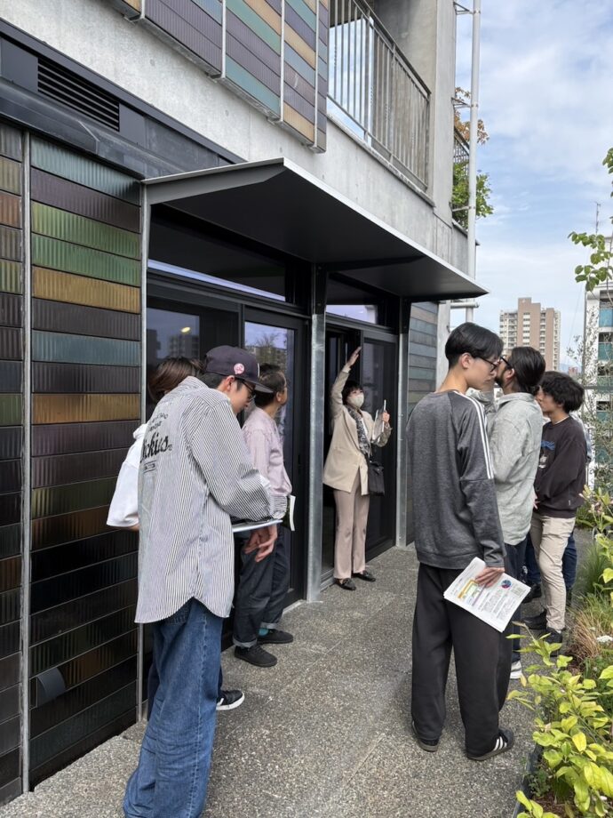 People lined up outside a modern building with glass doors and a dark metal canopy; some hold papers, and a masked person is near the doorway.