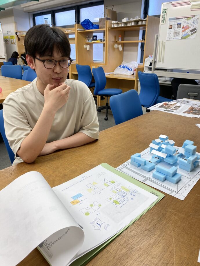 Student with glasses sits at a wooden table in a classroom, examining blue foam blocks and a diagram-filled notebook.