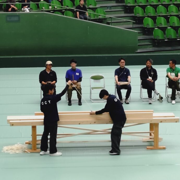 Judges seated in a gymnasium with green bleachers, two people standing and exchanging wooden planks while others sit nearby.