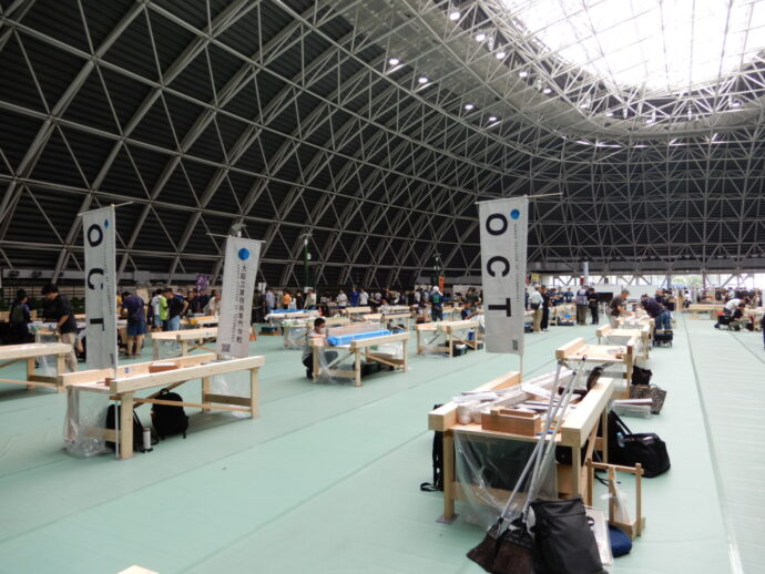 Wide view of a large indoor workshop hall with rows of workbenches and people at tasks under a lattice roof.