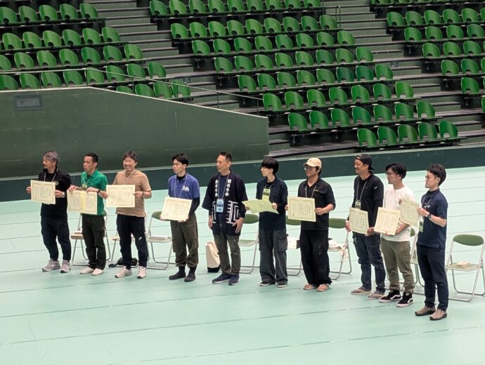 Group of people standing in a gym, each holding a certificate, posing for a photo with green stadium seats in the background
